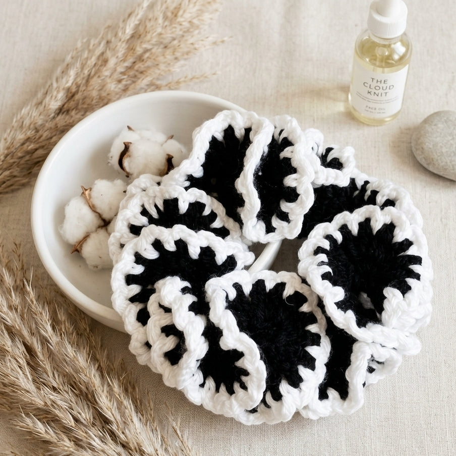 A set of handmade black and white crocheted hair scrunchies arranged in a white bowl with cotton bolls, dried pampas grass, and a small bottle of oil on a neutral fabric background.