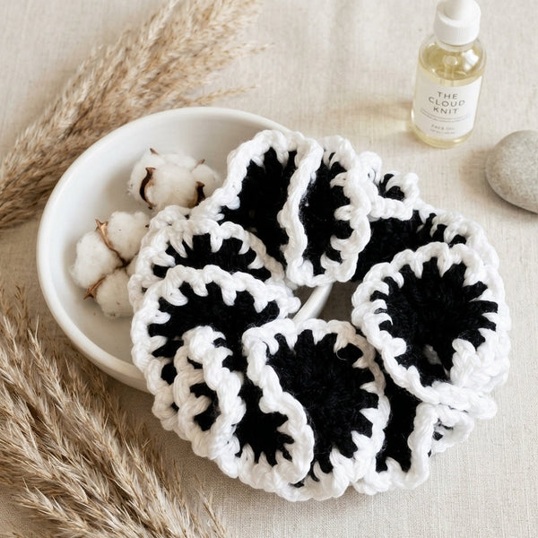 A set of handmade black and white crocheted hair scrunchies arranged in a white bowl with cotton bolls, dried pampas grass, and a small bottle of oil on a neutral fabric background.